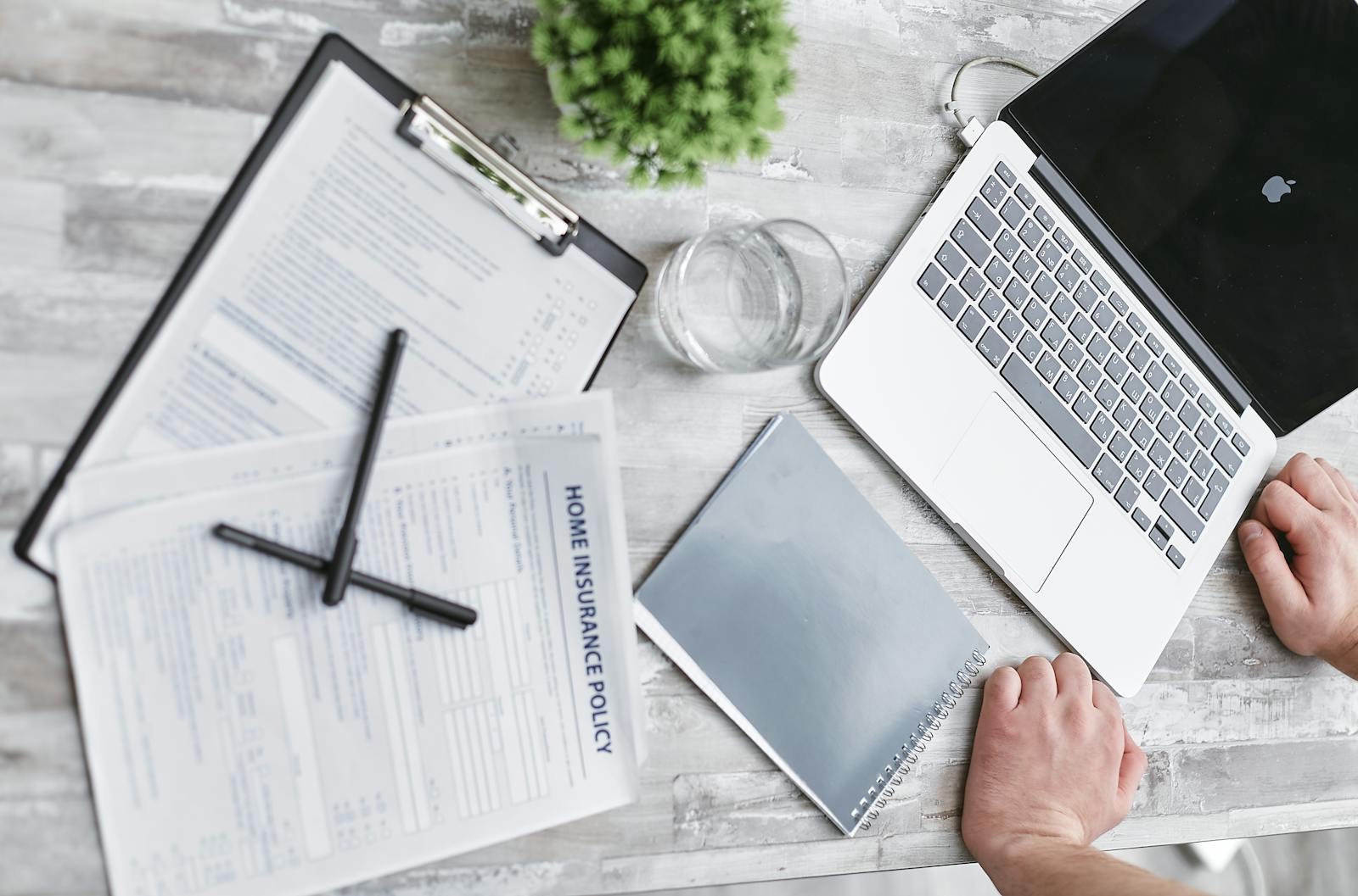 Top-down view of insurance claim forms, a laptop, and reading glasses on a broker's desk during a multilingual intake session.
