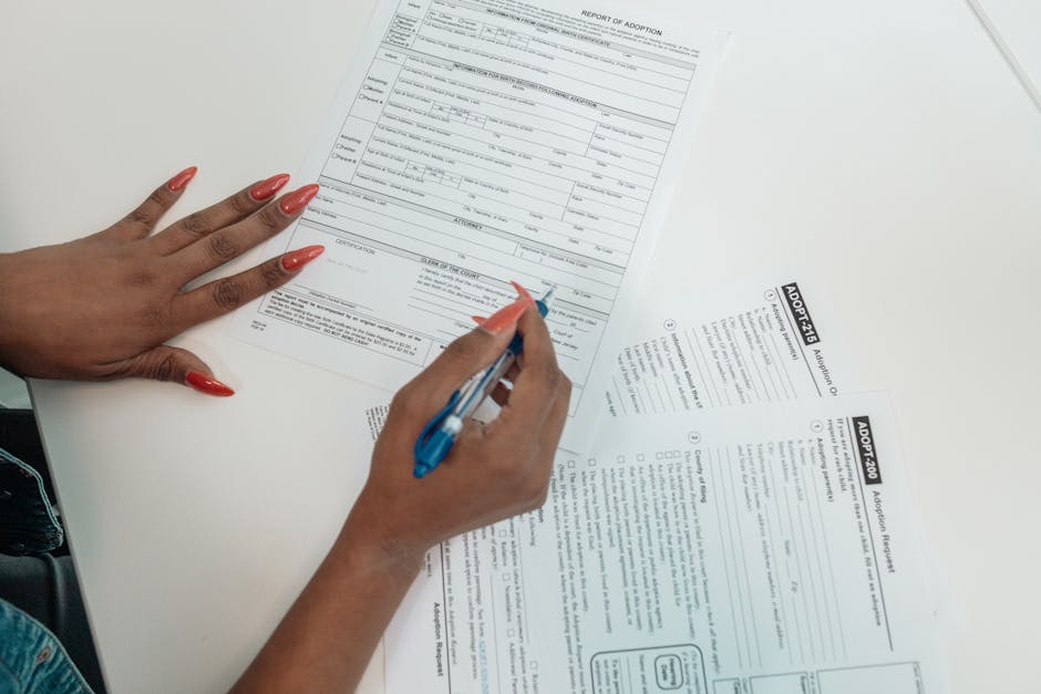 A close-up of a hand with a red manicure filling out a multi-page immigration application form on a wooden desk.
